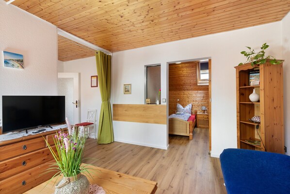 A room with a wooden ceiling, a television on a wooden dresser, a bed with a white headboard, a blue chair, and a wooden bookshelf.