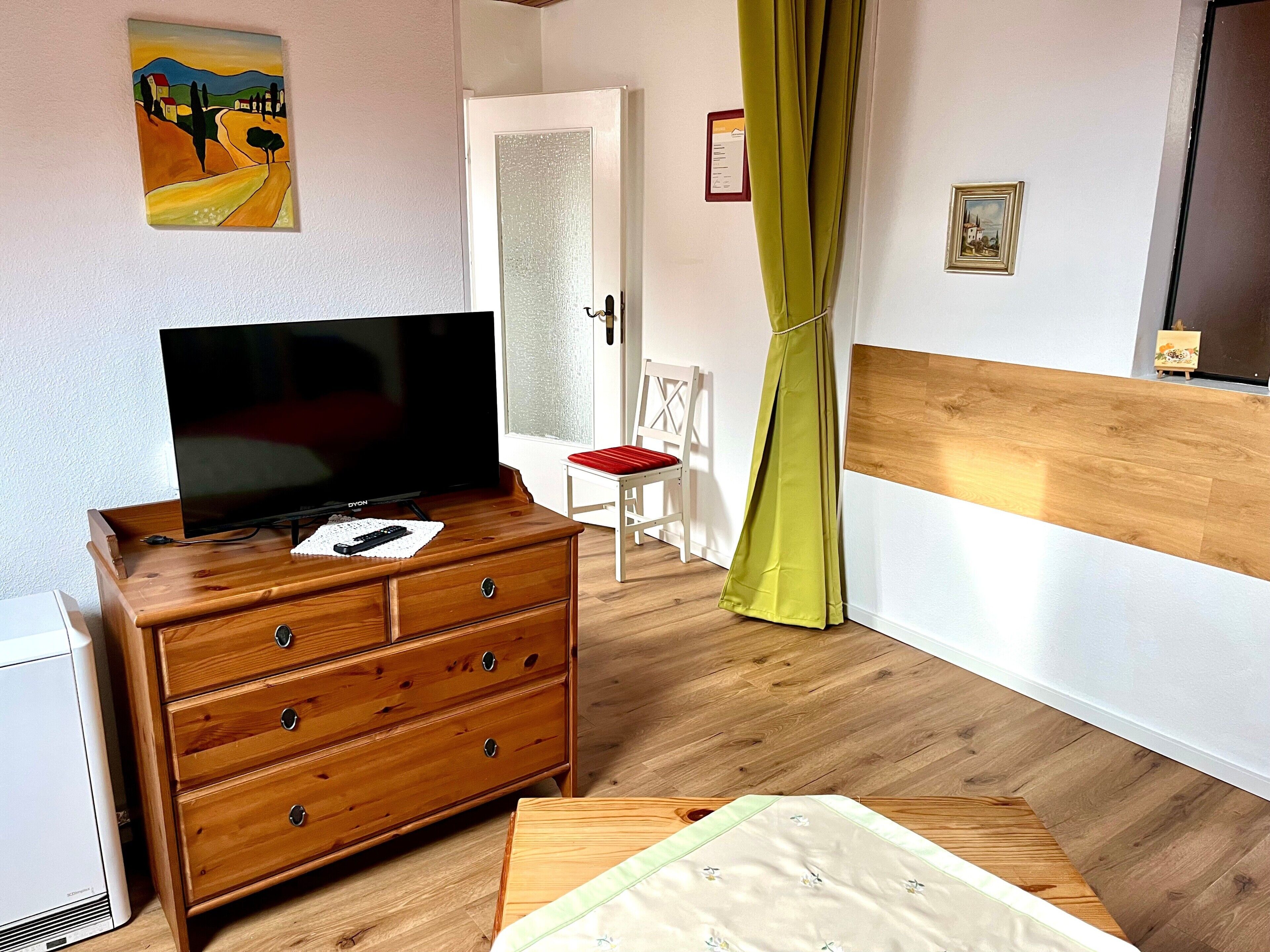Une pièce comprenant une commode en bois, un téléviseur, un lit avec une courtepointe, une porte et un tableau accroché au mur.