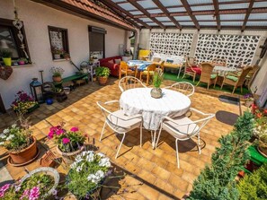 A covered patio with a round table and chairs, surrounded by potted plants and a dining area with chairs and a table.
