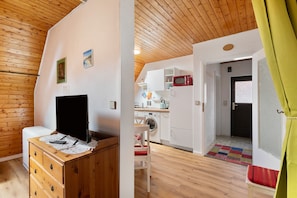 Une pièce avec un plafond en bois, un téléviseur posé sur une commode en bois, un lave-linge et un coin cuisine équipé de placards et d’un four à micro-ondes.