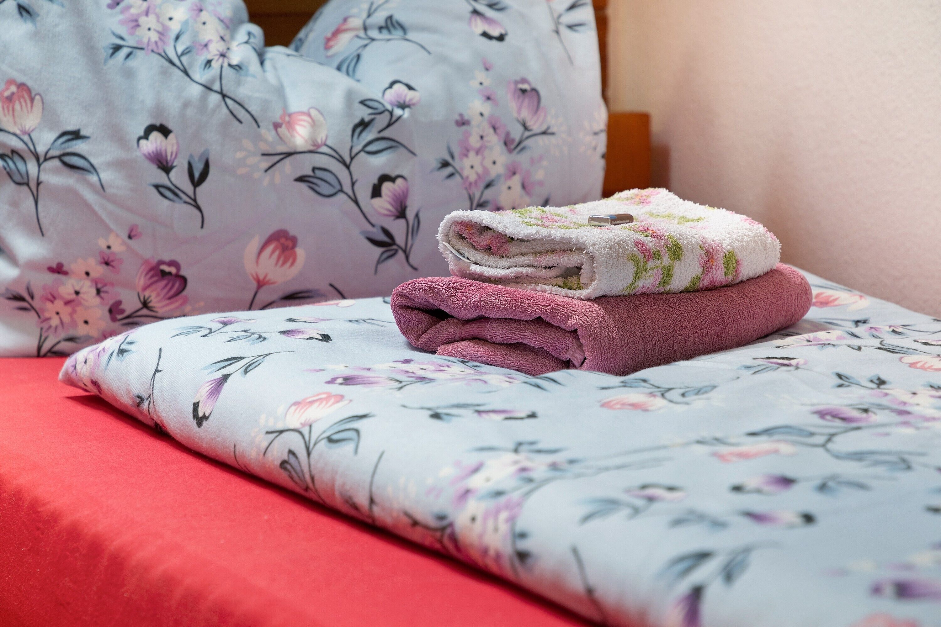 Una cama con ropa de cama floral, una manta roja y una pila de toallas dobladas.