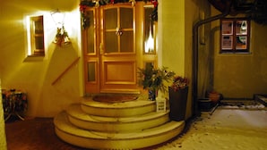 Un escalier menant à une porte ornée d’une couronne de Noël, une petite table recouverte d’une nappe à motifs floraux et un sol couvert de neige.