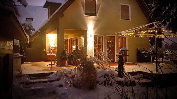 Une maison dont la cour est recouverte de neige, avec une véranda dotée de chaises et une guirlande lumineuse.