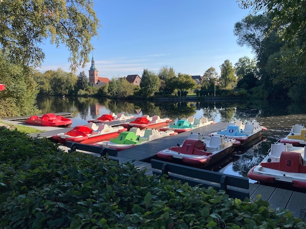 Ein ruhiger Fluss mit vertäuten Booten am Ufer, umgeben von Grün und einem Gebäude in der Ferne.