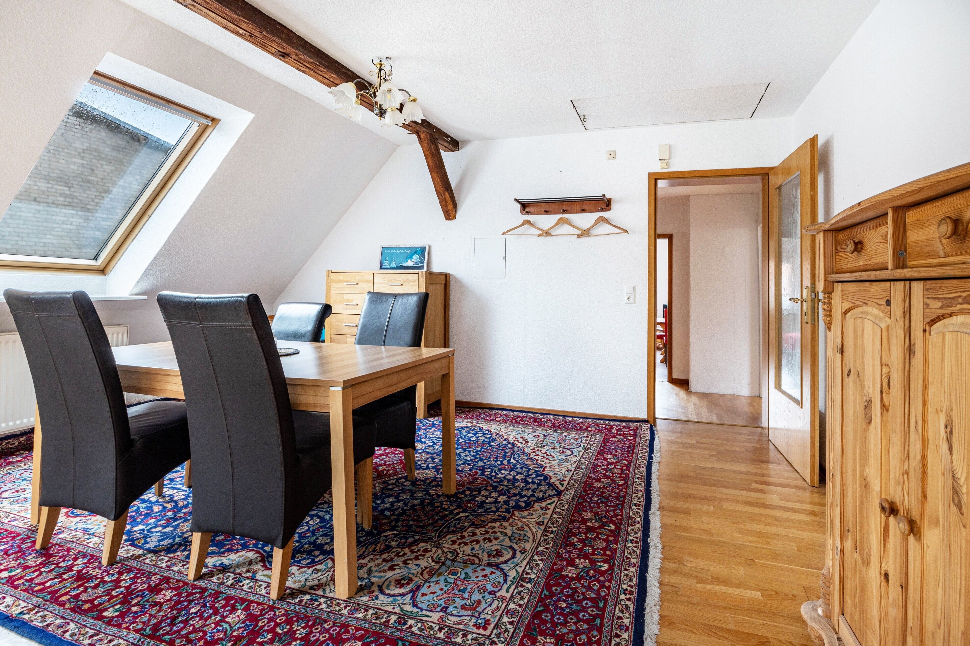 A dining area with a wooden table, black chairs, a patterned rug, a wooden cabinet, and a skylight.