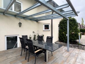 A covered patio with a glass table and chairs, a potted plant, and a wall-mounted light fixture. - Apartment 'Ferienwohnung 2' with Shared Garden, Balcony and Wi-Fi (Wasserburg (Bodensee))
