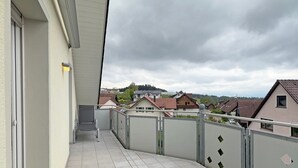 Uma varanda com piso de azulejos, grades de metal e vista para casas ao fundo.