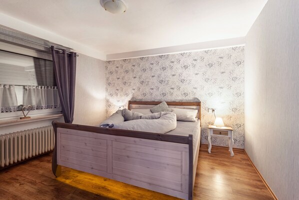 A bedroom with a wooden bed, a bedside table with a lamp, a radiator, and a window with curtains. - Vacation apartment "Wilde Schanze", 55sqm, with garden and wifi. (Brilon)