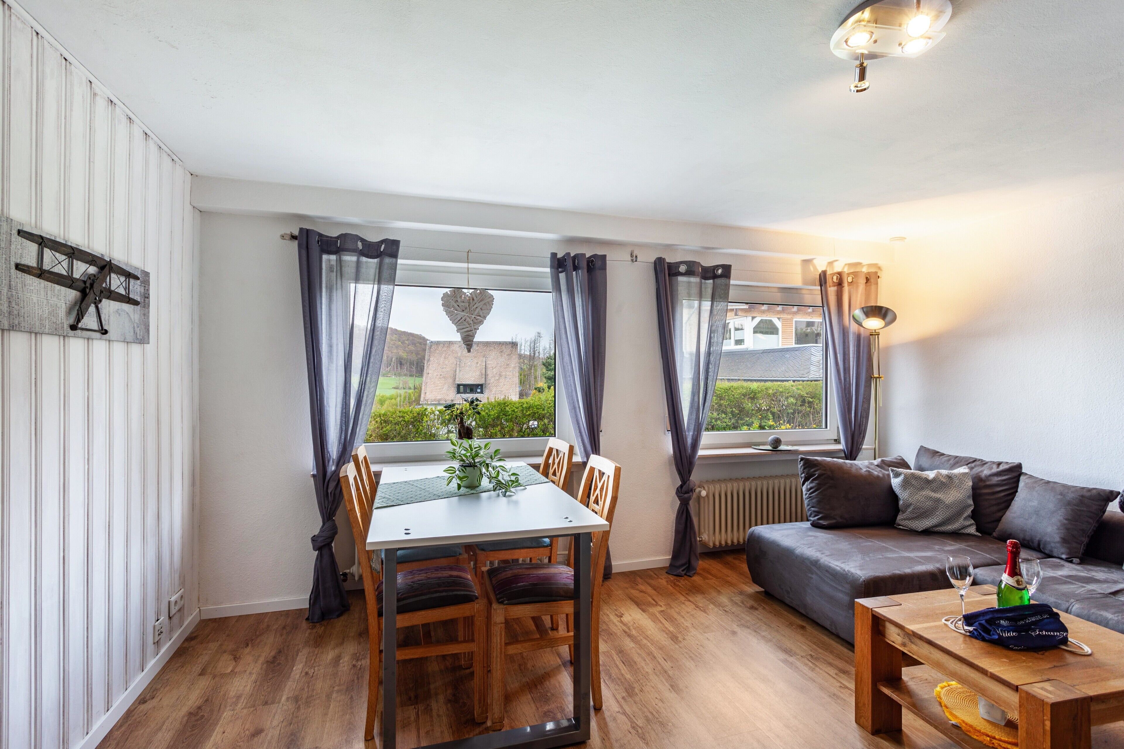 A room with a dining table, chairs, a sofa, a window with curtains, a hanging light fixture, and a radiator.