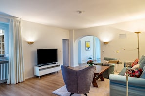 A modern living room with a flat-screen TV, a grey armchair, a blue sofa, a wooden coffee table, and a floor lamp.