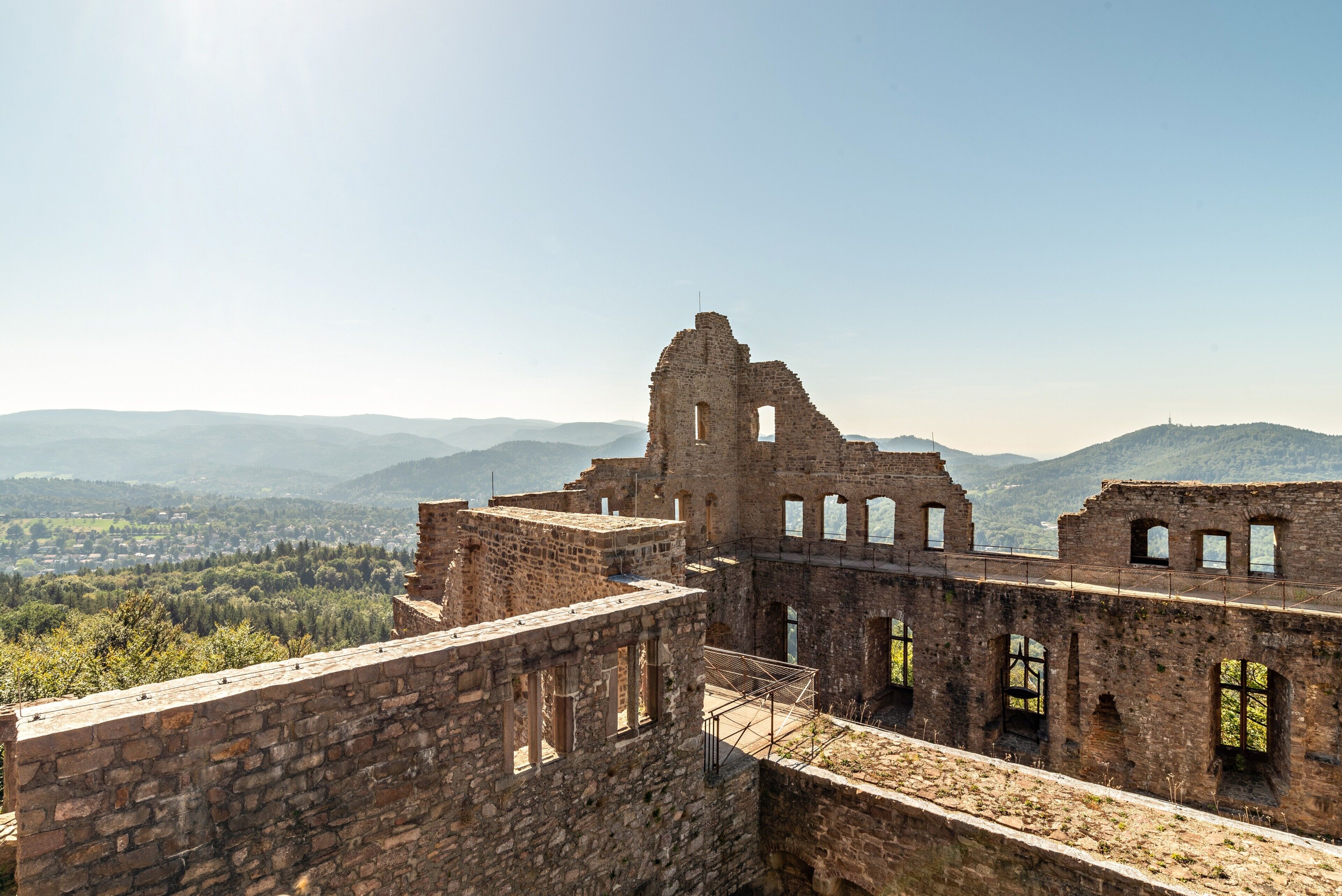 A historical stone structure with multiple levels and arched windows, overlooking a valley and distant mountains.