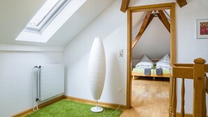 Una habitación con tragaluz, un radiador, una lámpara de pie, una cama con almohadas y una silla de madera.