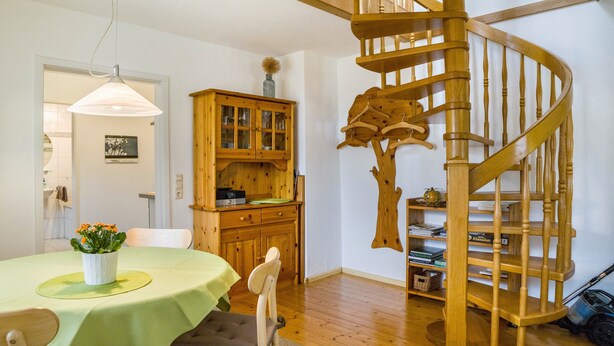 Una escalera de caracol con barandillas de madera, un comedor con una mesa y sillas verdes, y un armario de madera con puertas de vidrio.
