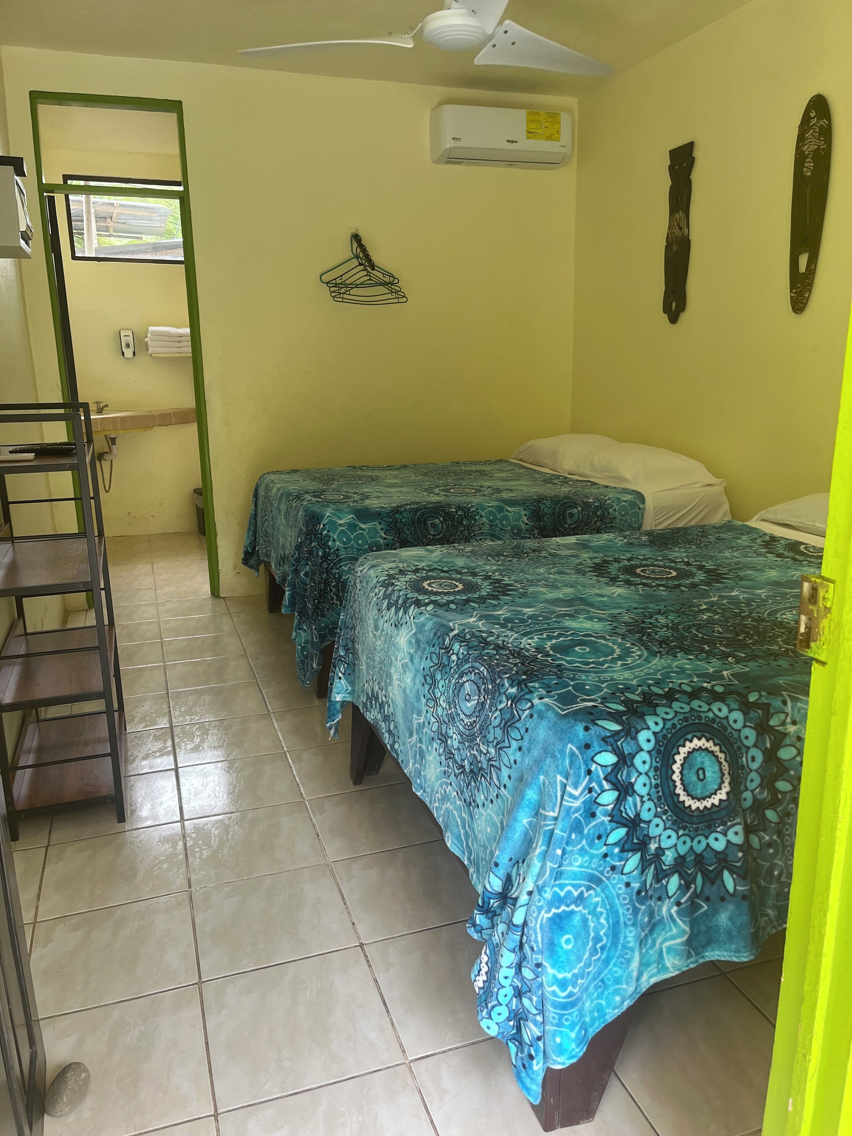 A room with two beds, a ceiling fan, an air conditioning unit, a small window, and a tiled floor.