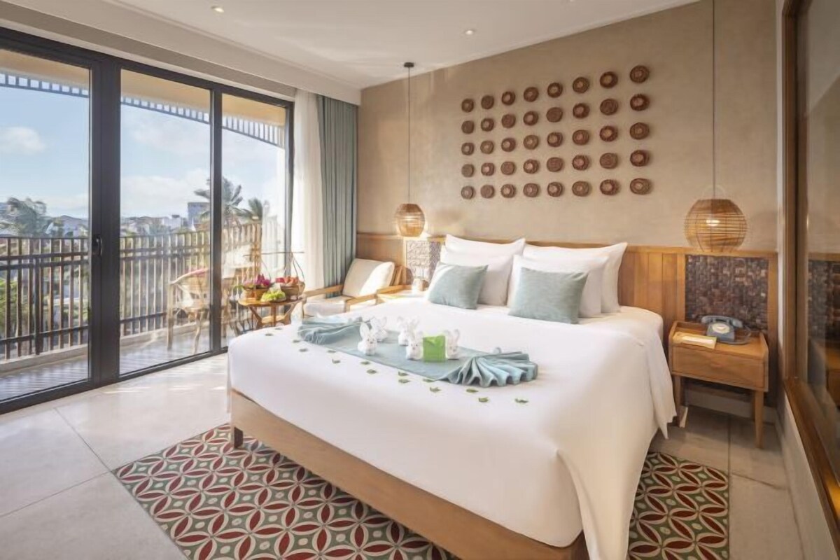Une chambre d’hôtel avec un grand lit, un balcon avec vue, une horloge murale et une petite table avec une lampe.