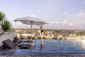 Ein Dachpool mit Infinity-Kante und Blick auf eine Stadtlandschaft, eine Lounge-Bereich mit Stühlen und einem Tisch sowie ein großer Sonnenschirm.