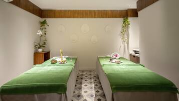 A spa room with two treatment tables, each covered with a green towel, and a wooden panel wall with decorative wall art.