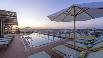 Un área de piscina en la azotea con sillones de descanso y vistas a la ciudad al atardecer.