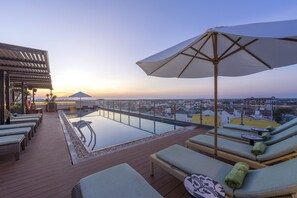 Un área de piscina en la azotea con sillones de descanso y vistas a la ciudad al atardecer.
