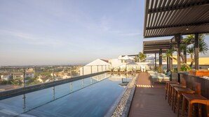 A rooftop infinity pool with a clear view of the cityscape, lounge chairs, and a covered seating area.