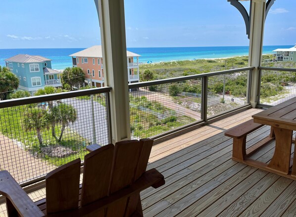 A wooden deck with chairs and a table, overlooking a beach and houses.