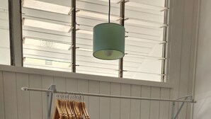 Een kamer met een bed, een houten bankje, een kledingrek, een prullenbak en een raam met jaloezieën.