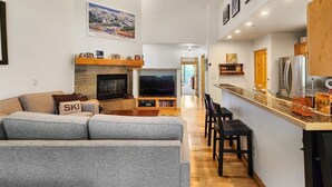 A modern living room with a gray sofa, a fireplace, a TV, and a kitchen area with a refrigerator and wooden cabinets. - Updated 3BD-3 Blocks to Gondola! (Breckenridge)
