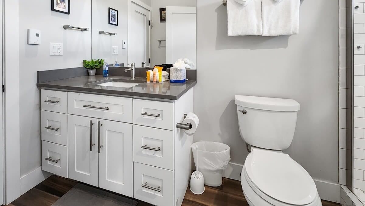 Un baño moderno con un lavamanos, encimera y inodoro blancos. Hay armarios con cajones, un cubo de basura y un espejo sobre el lavamanos.