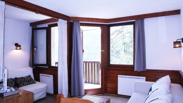 A cozy room with a wooden ceiling, a white sofa, a white armchair, a small table with a lamp, and a large window with curtains. - LARGE STUDIO 6 PEOPLE RESORT CENTER (LES ORRES 1650)