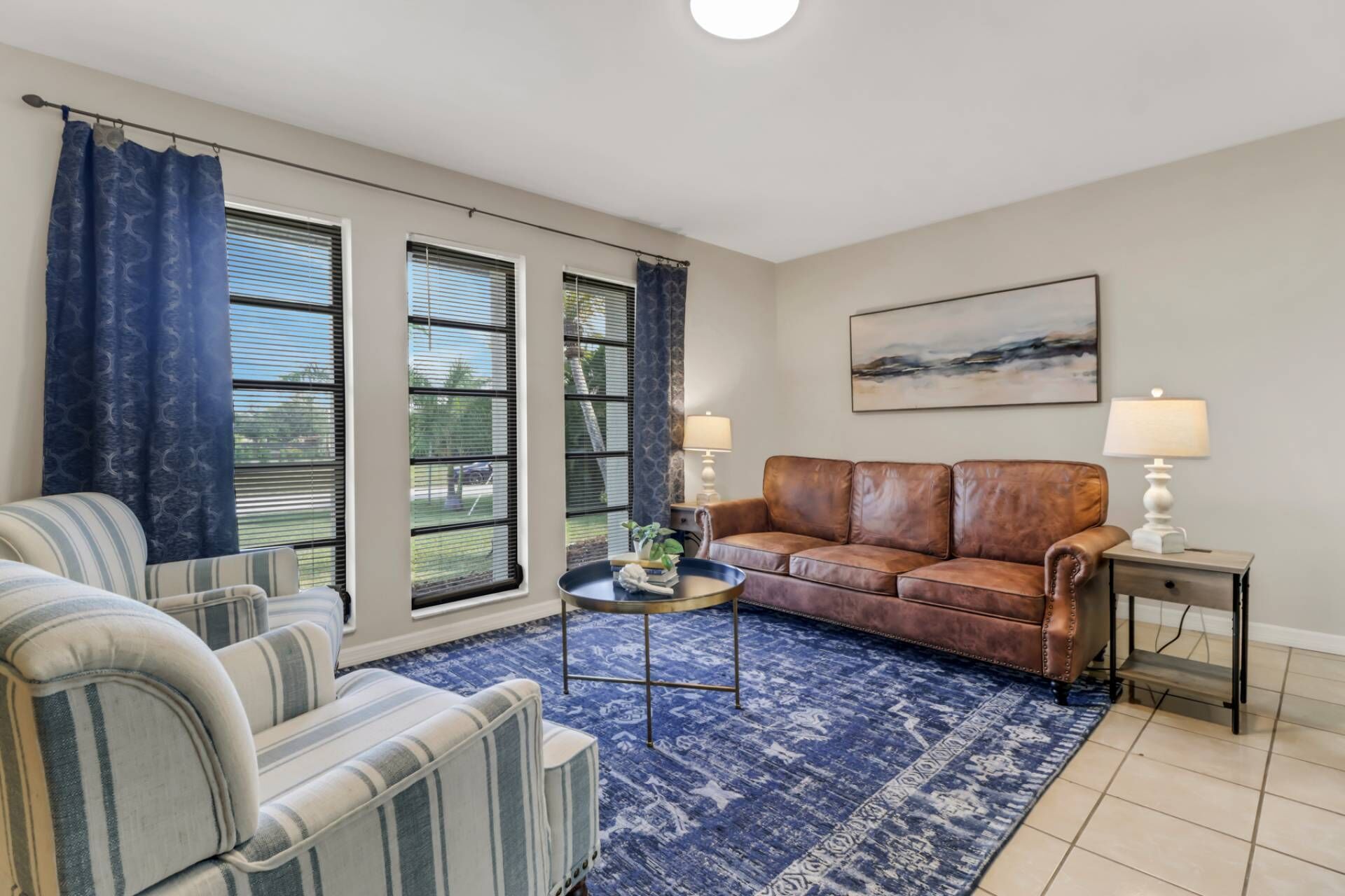 A living room with a brown leather sofa, striped armchairs, a blue patterned rug, and a painting on the wall.