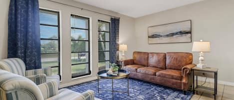 A living room with a brown leather sofa, striped armchairs, a blue patterned rug, and a painting on the wall.