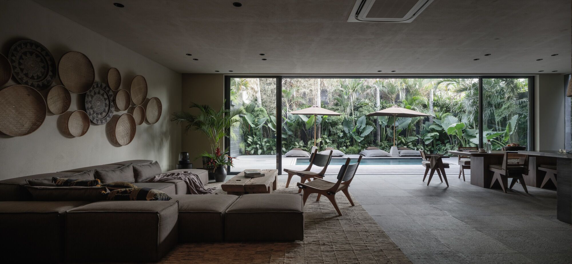 Uma sala de estar moderna com sofá, mesa de centro e cadeiras. Há uma janela grande com vista para um jardim e área de estar externa.