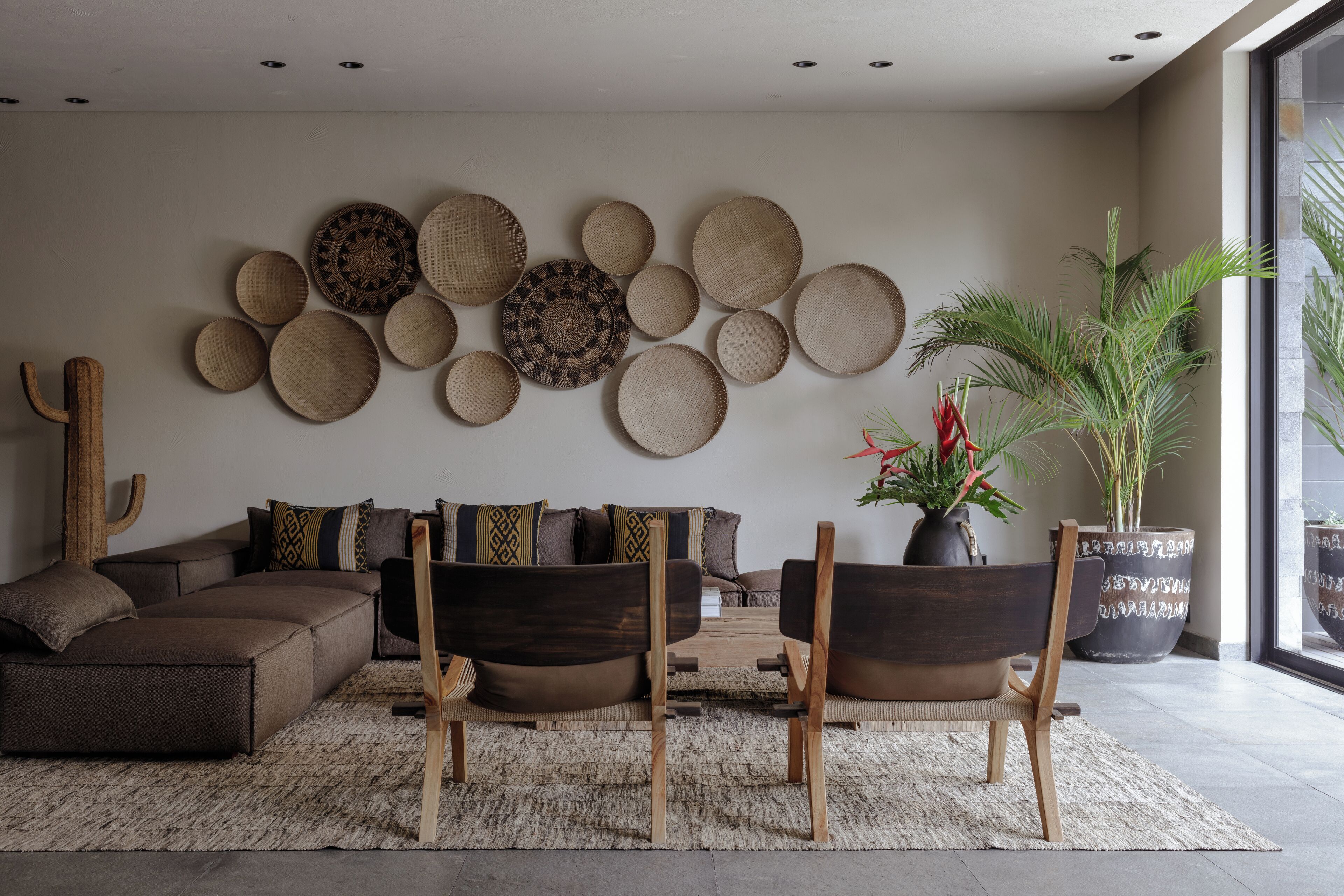 Un salon moderne avec un canapé d’angle, deux fauteuils en bois, une grande plante en pot et un mur décoré de tapis ronds tissés.