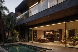 Uma casa moderna com piscina, área de refeições ao ar livre e cozinha visível através de grandes portas de vidro.