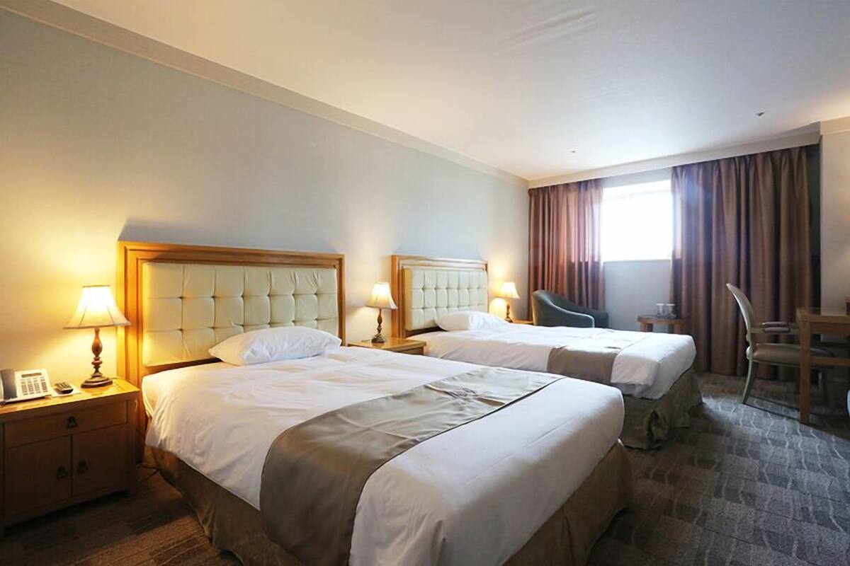Quarto de hotel com duas camas, uma escrivaninha, uma cadeira e uma janela com cortinas.