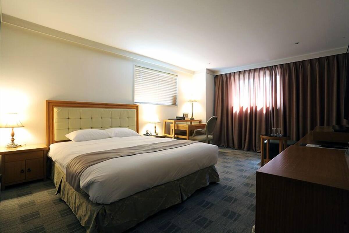 Quarto de hotel com uma cama grande, uma escrivaninha, uma cadeira e uma mesa pequena.