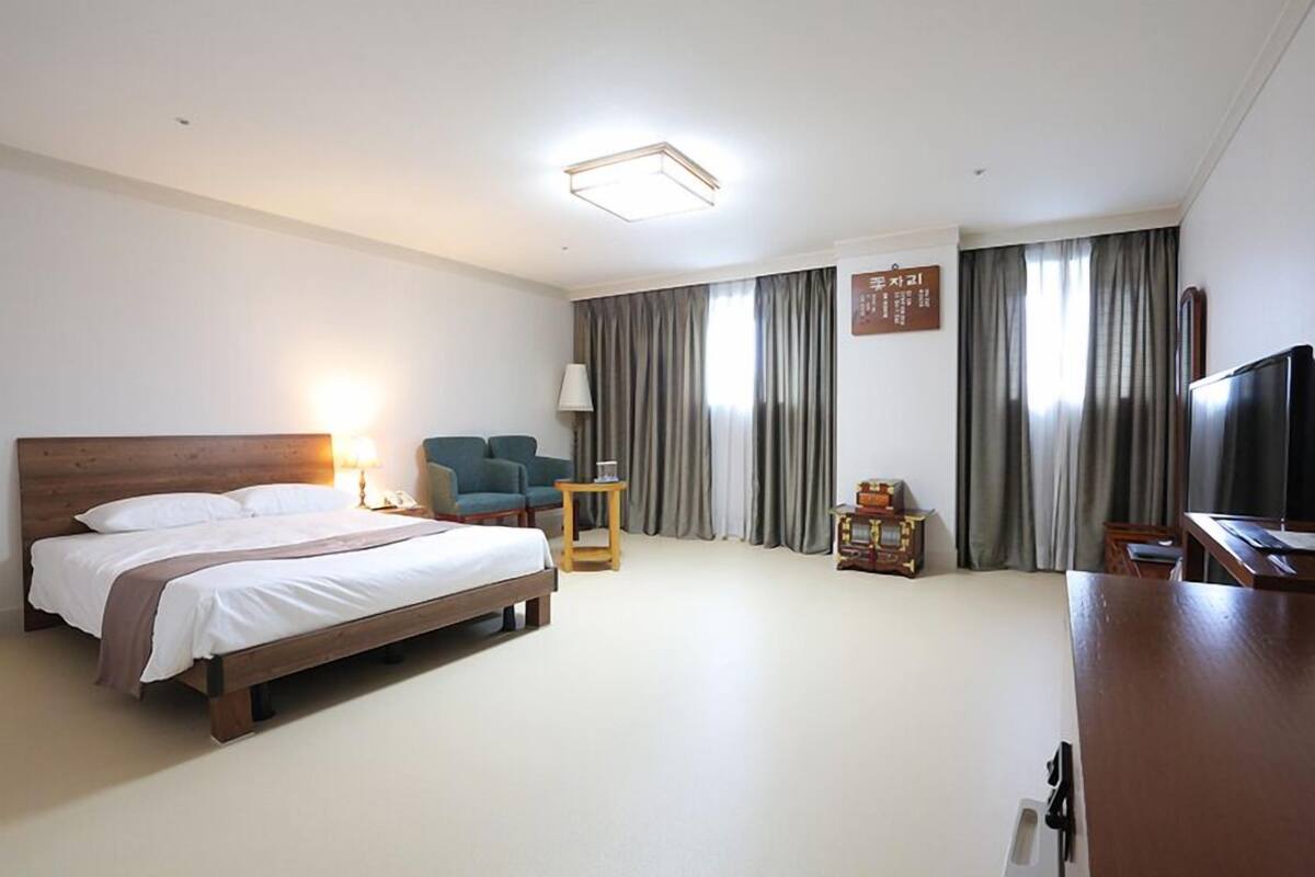 Quarto de hotel com cama de madeira, escrivaninha, cadeira, televisão e cortinas.