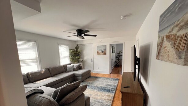 Un salon moderne avec un canapé d’angle, un ventilateur de plafond, un téléviseur à écran plat et un grand tableau accroché au mur.