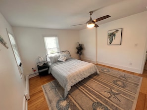 Une chambre à coucher avec un lit, un ventilateur de plafond, une fenêtre avec des stores, une table de chevet, une lampe, une plante et un tableau encadré au mur.