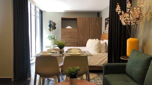 Moderni hotellihuone, jossa on lasinen ruokapöytä, vihreä sohva, valkoisilla tekstiileillä varustettu sänky ja suuri ikkuna, jossa on verhot.
