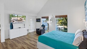 Un dormitorio con una cama, un tocador blanco, una silla, una ventana con vista a un cuerpo de agua y una puerta.
