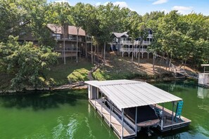 A dock with a covered seating area, a boat, and a house with a balcony overlooking a body of water. - Serenity Cove  - Margaritaville Home w/Private Dock! (Osage Beach)