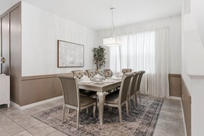 A dining room with a rectangular table, six chairs, a chandelier, a painting on the wall, and a plant.