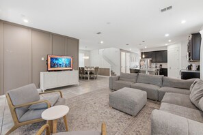 Ein modernes Wohnzimmer mit Flachbildfernseher, grauen Sofas, einem Essbereich mit Stühlen und einer Küche im Hintergrund.