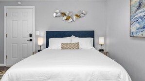A neatly arranged bedroom with a large bed, two bedside tables with lamps, a decorative wall piece above the bed, and a door to the left. - Cozy Studio in the Heart of Downtown Kimmswick (Imperial)