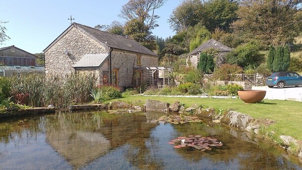A stone building with a pond in front, surrounded by greenery and a parked car.