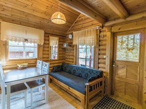 L’intérieur d’une cabane en rondins, avec une table à manger, des chaises, un canapé, un luminaire suspendu et une fenêtre ornée de rideaux en dentelle.