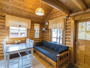 Interior de una cabaña de troncos con una mesa de comedor de madera, sillas, un banco con cojín floral y una lámpara colgante.