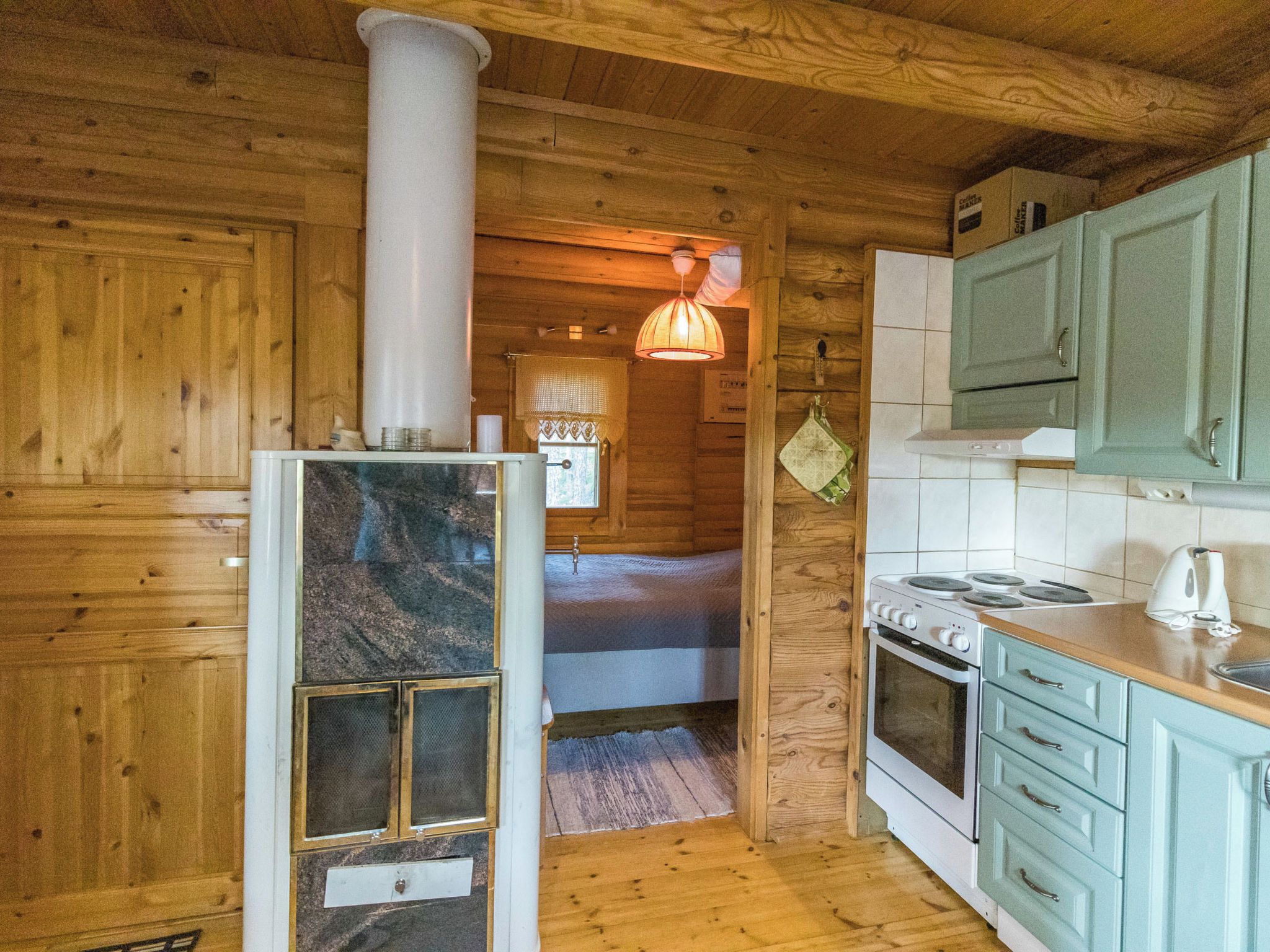 L’intérieur d’un chalet en bois comprend un coin cuisine équipé d’un poêle, de placards et d’une fenêtre avec un rideau.