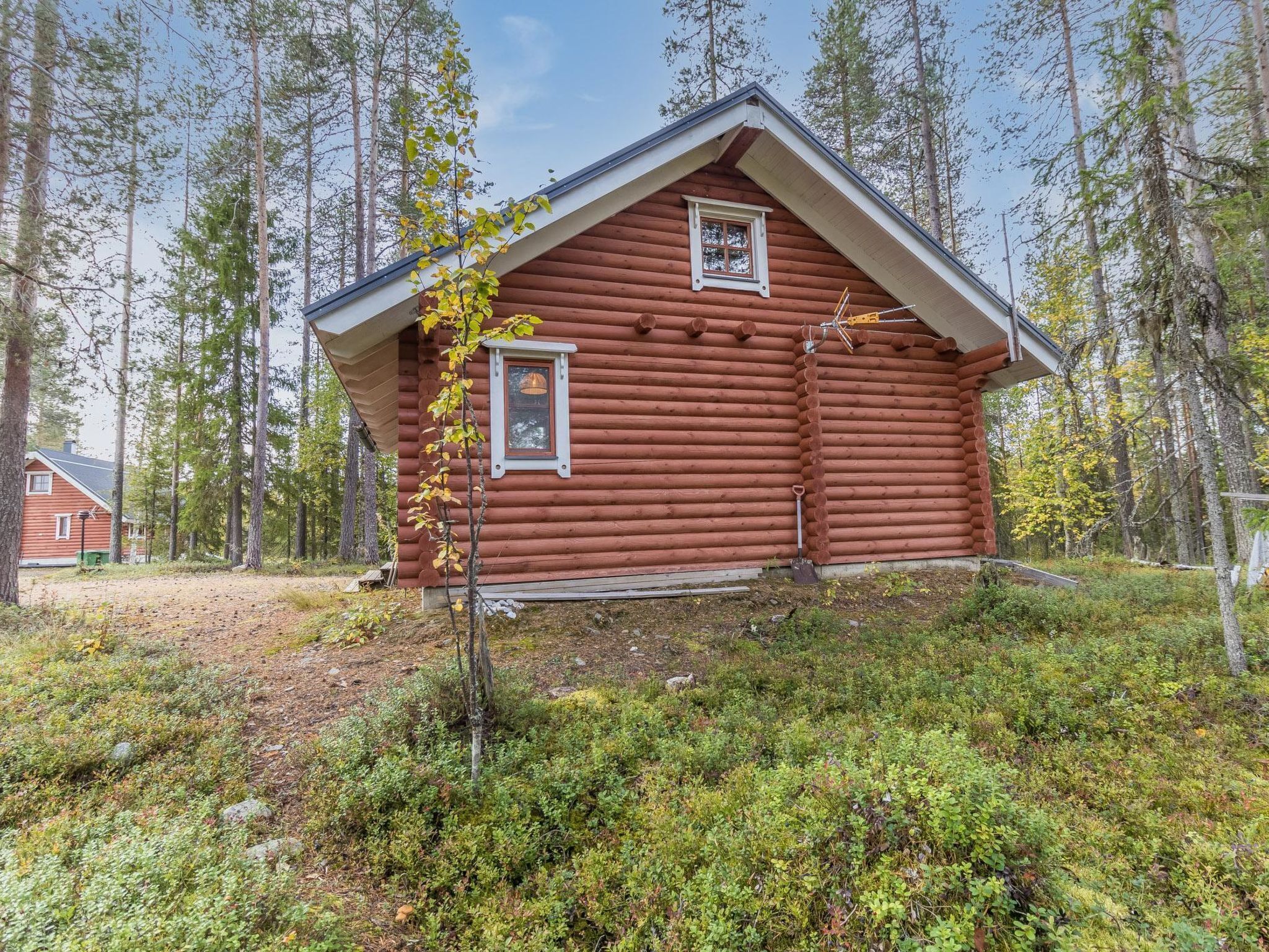 Hirsimökki, jossa on harjakatto ja kaksi ikkunaa, ja sen ympärillä on metsäinen alue.